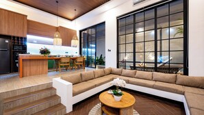 Une cuisine moderne avec un plafond en bois, un coin repas doté d’une grande fenêtre et un salon avec un canapé d’angle et une table basse ronde.