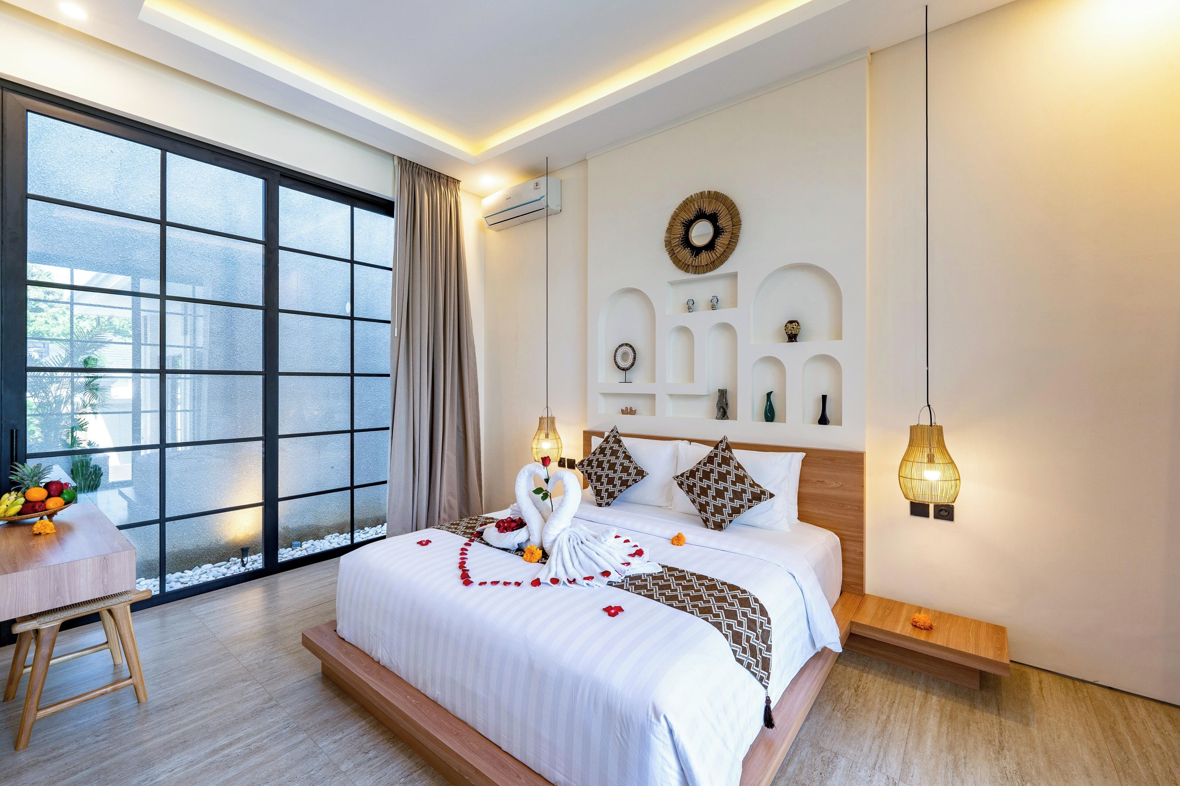 A modern hotel room with a large window, a bed with a decorative swan centerpiece, a small table with fruit, and a wall-mounted light fixture.