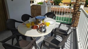 Une table ronde avec une bouilloire en métal, des bols jaunes et rouges, et une assiette blanche, le tout sur un balcon agrémenté de quatre chaises.