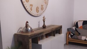 Un salon comprenant une grande horloge murale, une console en bois agrémentée d’objets décoratifs, un canapé et une plante en pot.