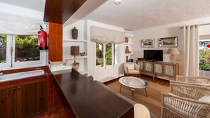 Ein modernes Wohnzimmer mit einer dunklen Holztheke, weißen Wänden und einem Fernseher. Es gibt Korbstühle, einen kleinen runden Tisch und einen an der Wand befestigten Feuerlöscher.