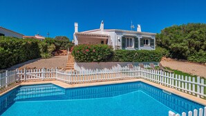 Ein Schwimmbecken mit weißem Zaun, ein Haus mit rotem Dach und üppiges Grün im Hintergrund.