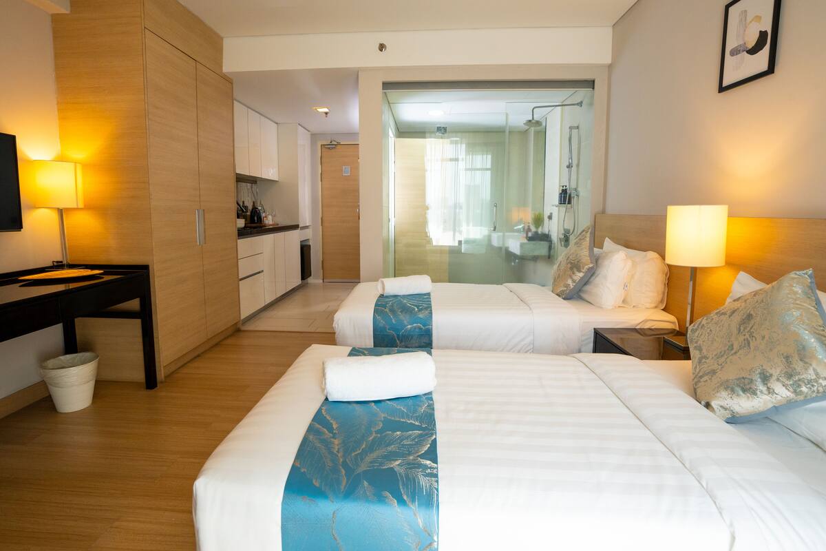 Ein Hotelzimmer mit zwei Betten, einem Schreibtisch, einer kleinen Küchenzeile und einem durch eine Glastür einsehbaren Badezimmer.