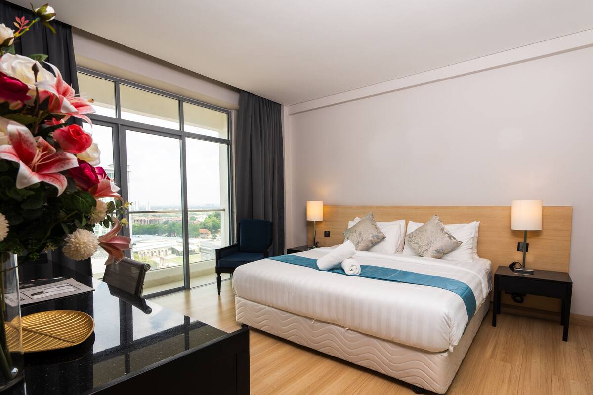 Ein Hotelzimmer mit einem großen Bett, zwei Nachttischen, einem Schreibtisch mit Stuhl sowie einer Blumenvase auf dem Schreibtisch und Blick auf die Stadt durch große Fenster.