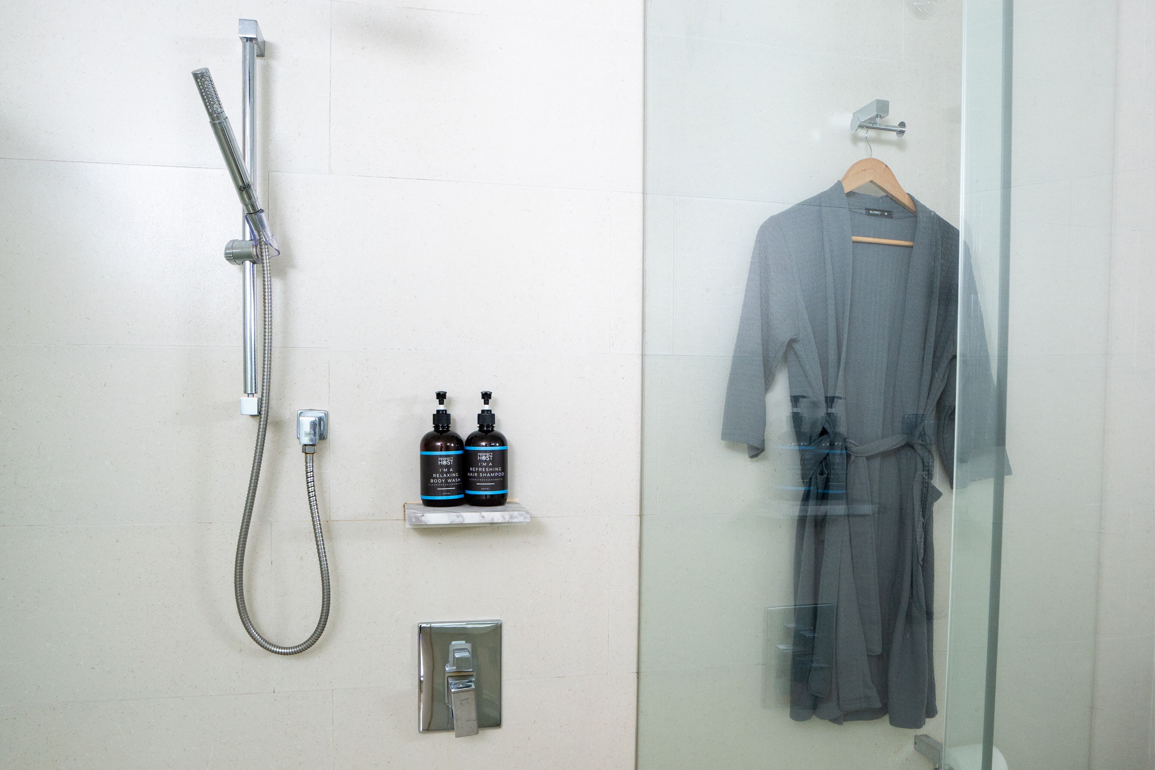 A bathroom with a shower, two shampoo bottles, a towel rack with a robe, and a towel dispenser.