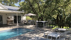 A pool area with lounge chairs and a table, surrounded by trees and a building. - Luxury 5 roomed Villa with A/C, swimming pool etc. in Victoria Falls, Zimbabwe (Victoria Falls)