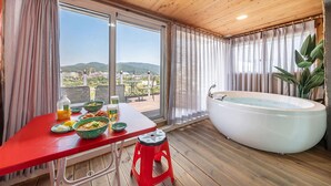 A room with a wooden ceiling, a large window with a view of a cityscape, a red table with bowls of food, a white bathtub, and a red stool. - Gyeongju Star spa Pension (Gyeongju)
