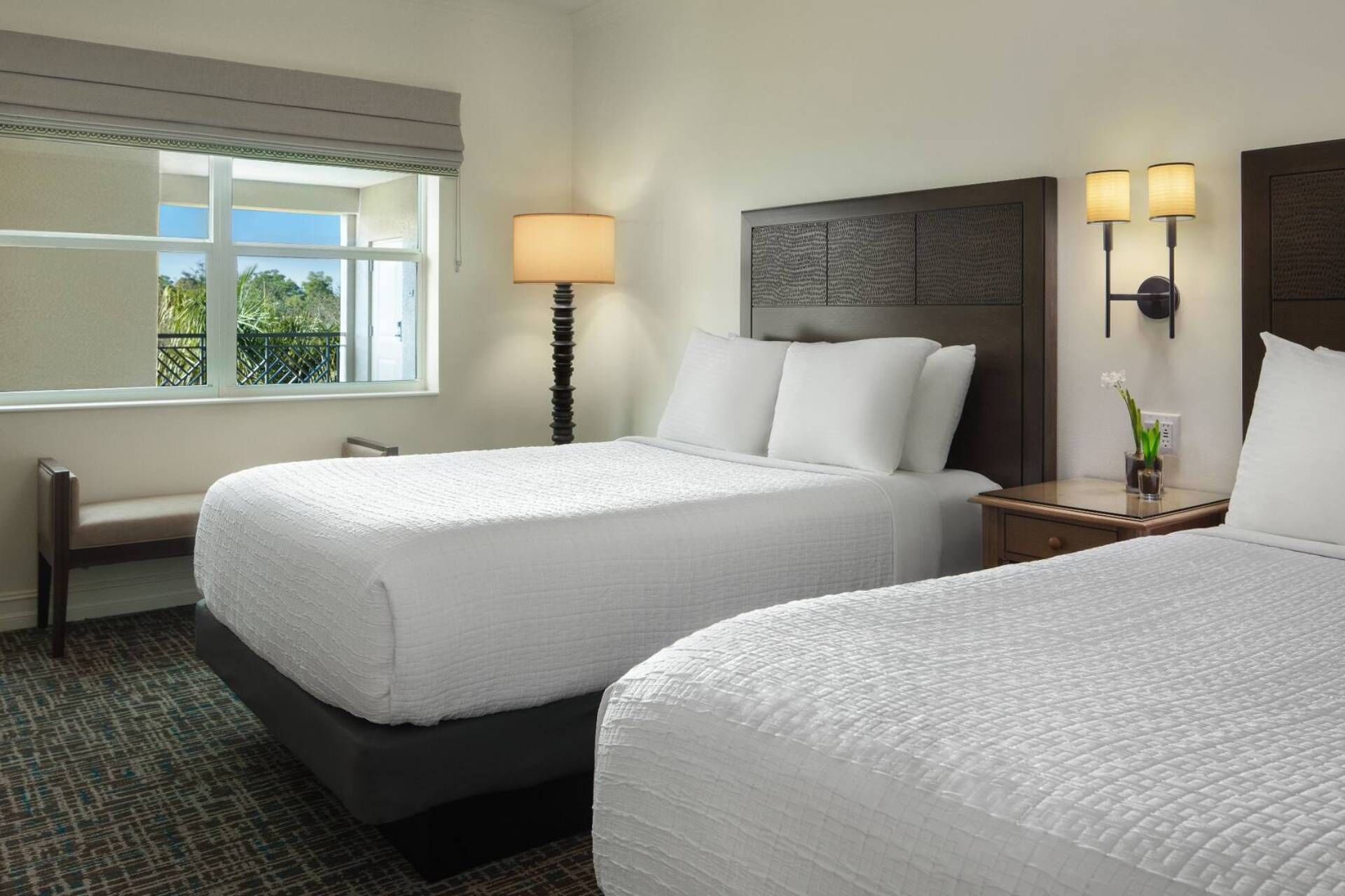 Quarto de hotel com duas camas, janela com vista, luminária e mesa de cabeceira com um vaso de flores.