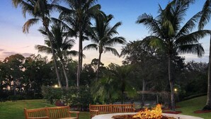 Un foyer circulaire en pierre, agrémenté d’un banc et d’un banc en bois, situé dans un jardin aménagé avec des palmiers.