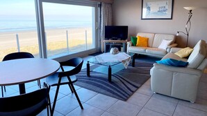 Una sala de estar con una mesa de vidrio, sillas, un sofá, un televisor de pantalla plana y vista a la playa.
