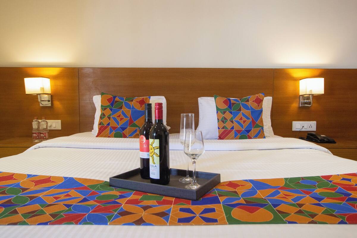 Quarto de hotel com cama bem arrumada, duas almofadas com padrões geométricos, bandeja com duas garrafas de vinho e um copo, e duas luminárias fixadas na parede.