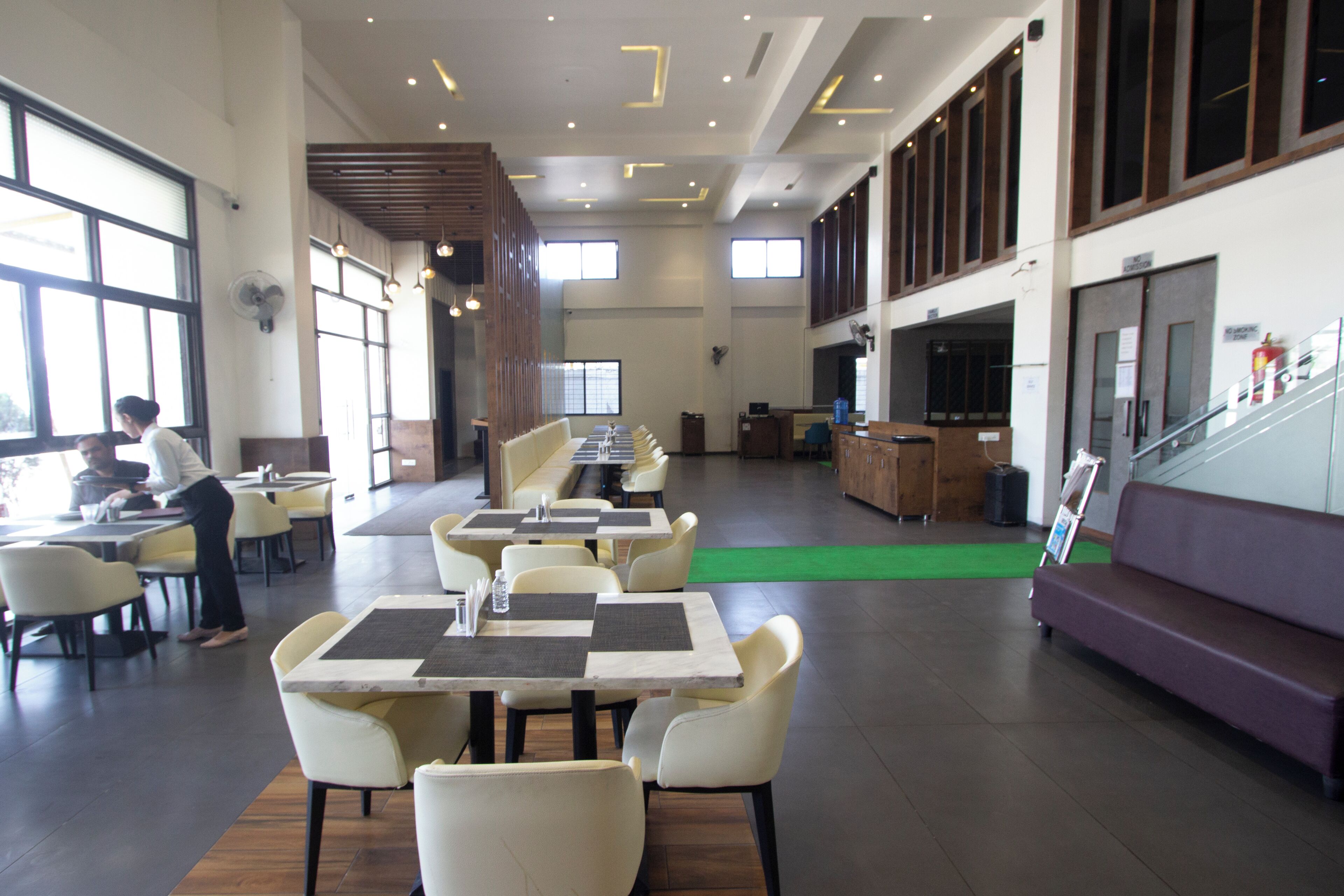A spacious indoor area with multiple tables and chairs arranged for dining. There are large windows allowing natural light, and a staircase is visible in the background.