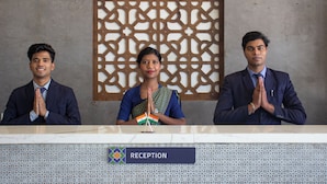 Three individuals seated at a reception desk with a 'RECEPTION' sign, a decorative wall, and a patterned backdrop. - Monarch Express (Rajgurunagar)