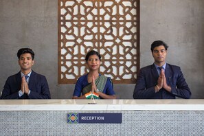 Three individuals seated at a reception desk with a 'RECEPTION' sign, a decorative wall, and a patterned backdrop. - Monarch Express (Rajgurunagar)