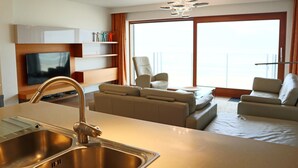 Habitación de hotel moderna con cocina, zona de estar con sofá y sillones, televisión y un amplio ventanal con vistas.