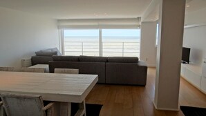 A modern living room with a large window overlooking the sea, a grey sofa, a wooden dining table, and a flat-screen TV mounted on the wall. - Nieuwpoort Beachfront Comfort (Nieuwpoort)