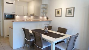Una cocina moderna con armarios blancos, horno integrado y comedor con mesa blanca y sillas de mimbre.