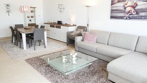 Een moderne woonkamer met een grijze bank, een glazen salontafel en een eethoek met een houten tafel en stoelen.