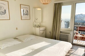A bedroom with a bed, a bathroom with a sink and mirror, and a view of buildings through a window. - Picturesque apartment in Nieuwpoort (Nieuwpoort)