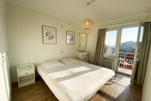 A bedroom with a bed, bedside table, nightstand, window with curtains, and a view of the city.
