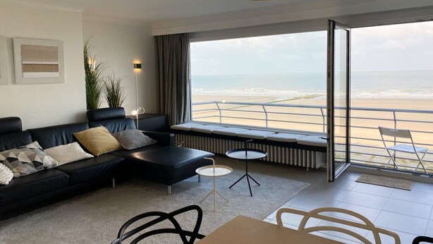A modern living room with a black sofa, a small round table, and a view of the beach through large windows. - Picturesque apartment in Nieuwpoort (Nieuwpoort)