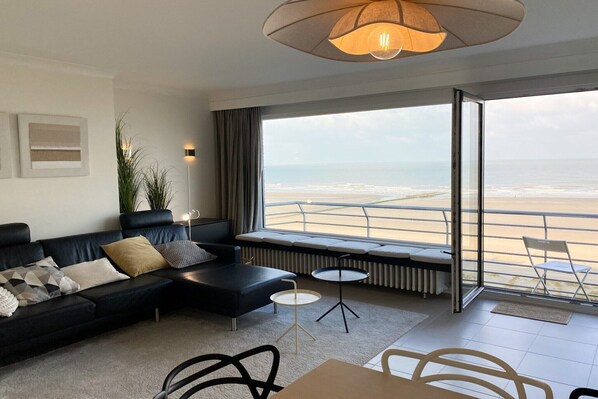 A modern living room with a black sofa, a small round table, and a view of the beach through large windows.