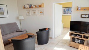 Uma sala de estar com um sofá cinzento, duas poltronas pretas, uma mesa de centro de madeira, um rack de TV com televisão de tela plana e uma estante com quadros e objetos decorativos.