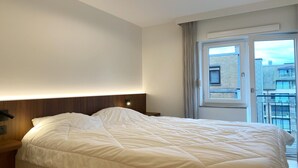 A hotel room with a large bed, a wooden headboard, a bedside table, and a window with a view of buildings.