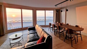 A spacious living area with a large window overlooking the sea, a leather sofa, a dining table with chairs, and a small coffee table.