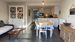 A modern living room with a sofa, coffee table, and dining area with chairs and a table. There is a kitchen in the background with cabinets and appliances. - Apartment in Nieuwpoort near Sandy Beaches (Nieuwpoort)