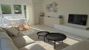 Ein modernes Wohnzimmer mit einer Couch, zwei Couchtischen, einem Essbereich mit Stühlen, einer Küchenzeile und einem an der Wand montierten Fernseher.