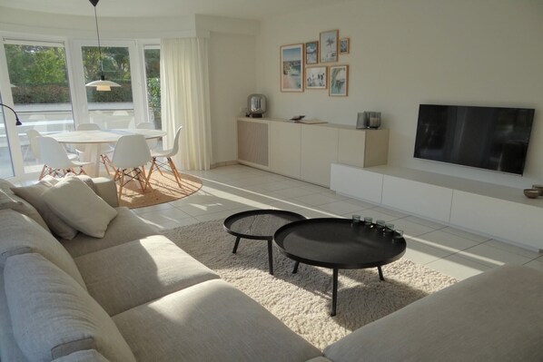 A modern living room with a sofa, two coffee tables, a dining area with chairs, a kitchen area, and a TV mounted on the wall. - Apartment in Nieuwpoort with Sea Views (Nieuwpoort)
