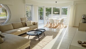 Ein modernes Wohnzimmer mit einem beigen Sofa, zwei runden Couchtischen und einem Essbereich mit einem weißen Tisch und Stühlen.
