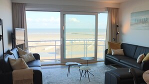 A modern hotel room with a large sliding glass door leading to a beach view, a black leather sofa, a small round table, and a flat-screen TV. - Apartment in Niewpoort near the Seaside (Nieuwpoort)