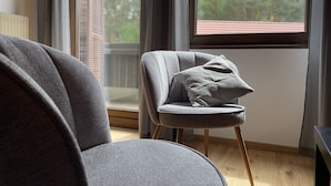 Uma sala de estar moderna com um sofá e uma poltrona cinzas, uma mesinha lateral e uma janela com vista para árvores e um edifício.