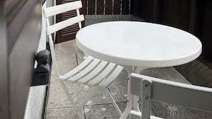 Um pátio com uma mesa e cadeiras brancas, cerca de madeira, e vista para árvores e uma casa ao fundo.