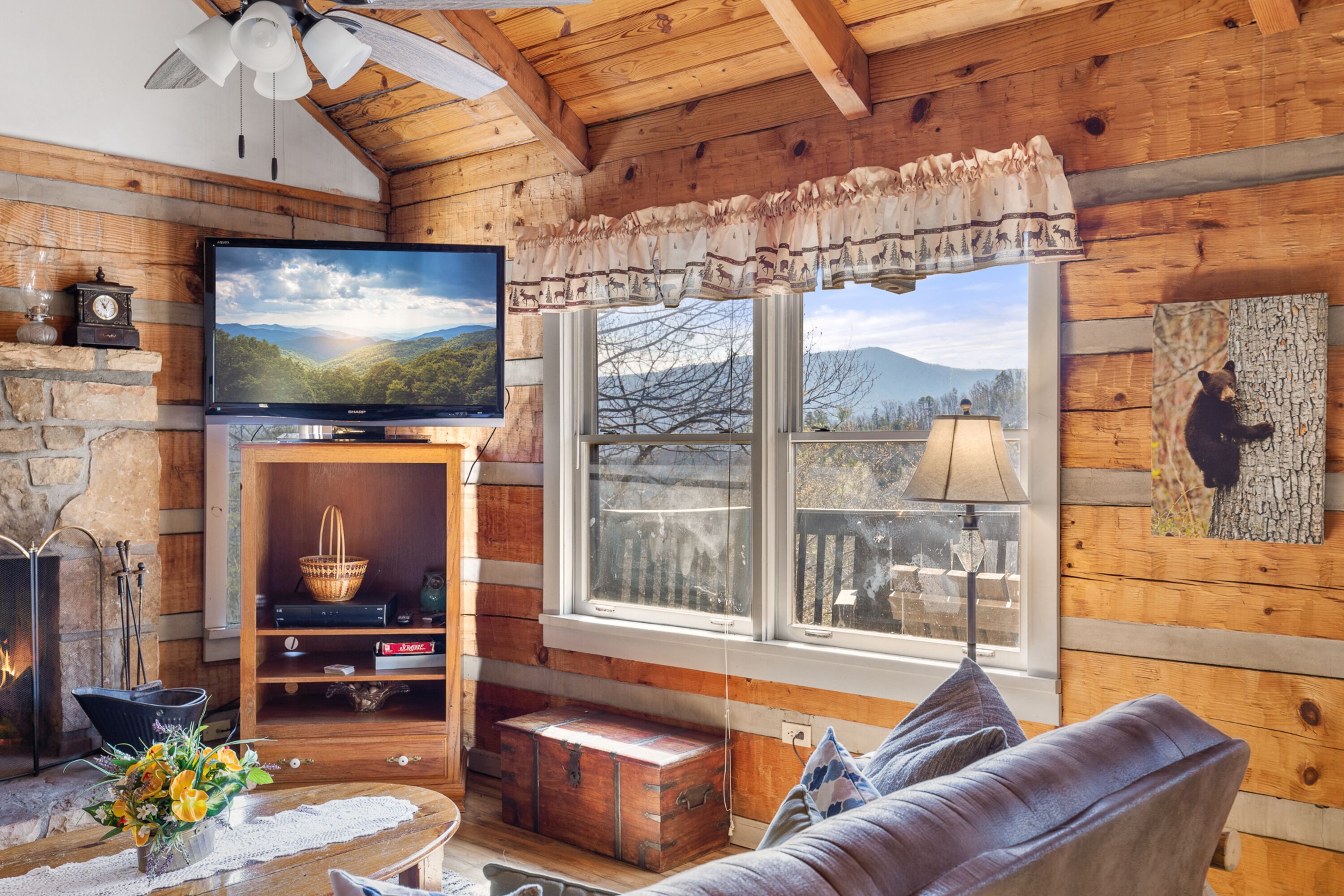 A cozy cabin interior with a fireplace, a television mounted on a wooden wall, a large window with a view of the outdoors, and a bear-themed artwork on the wall.