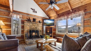 A log cabin interior with a stone fireplace, wooden furniture, and a large window offering a view of the outdoors. - Mountain Vintage (Sevierville)