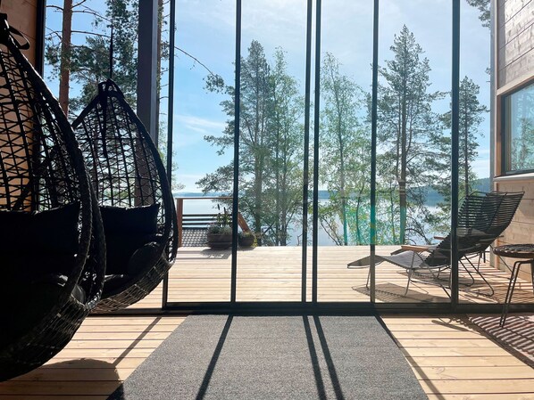 A wooden deck with outdoor furniture, including a hanging chair and lounge chairs, overlooking a forest and a body of water.