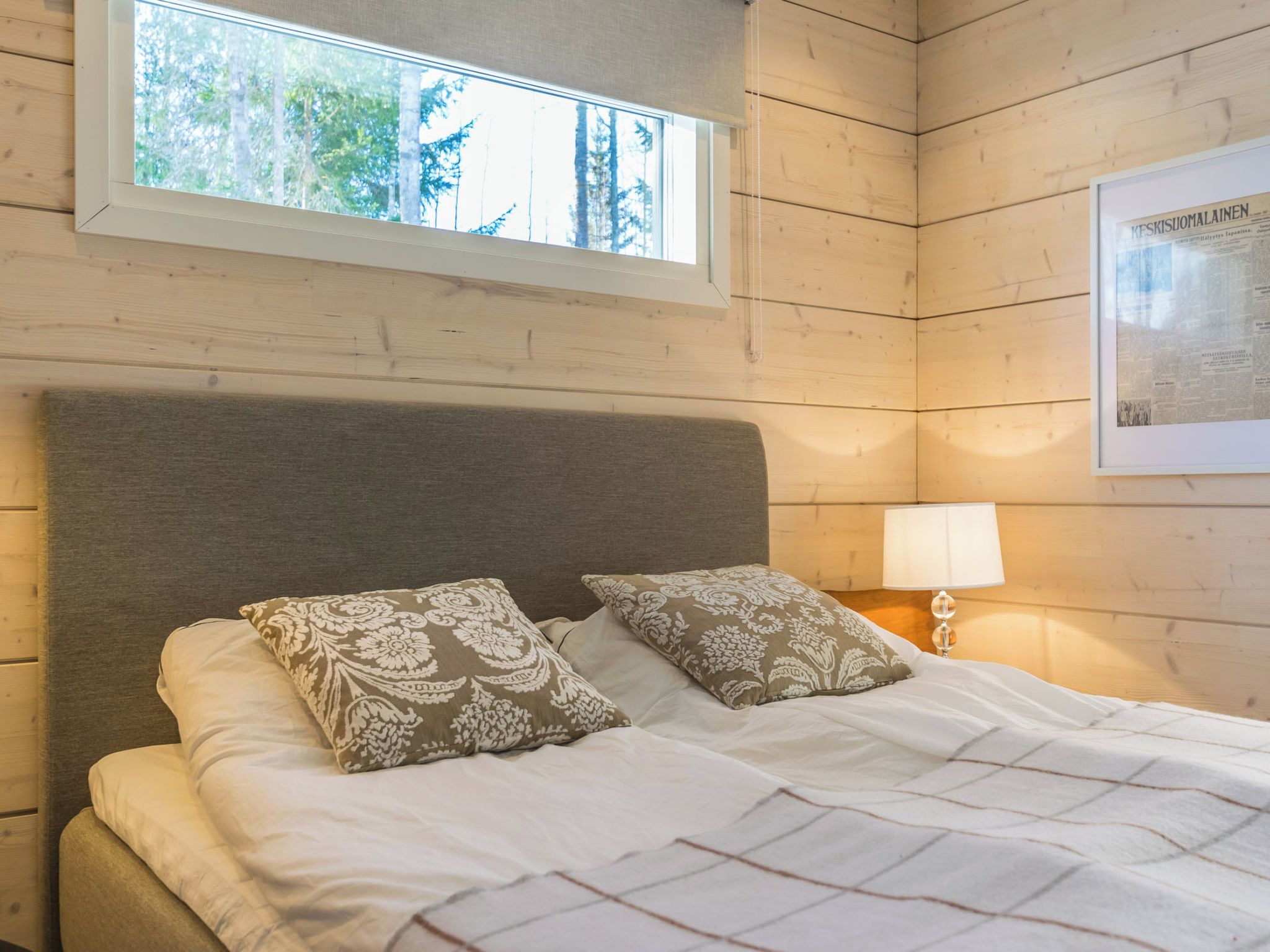 A bedroom with a bed, two patterned pillows, a nightstand with a lamp, a window with a view of trees, and a framed newspaper on the wall.