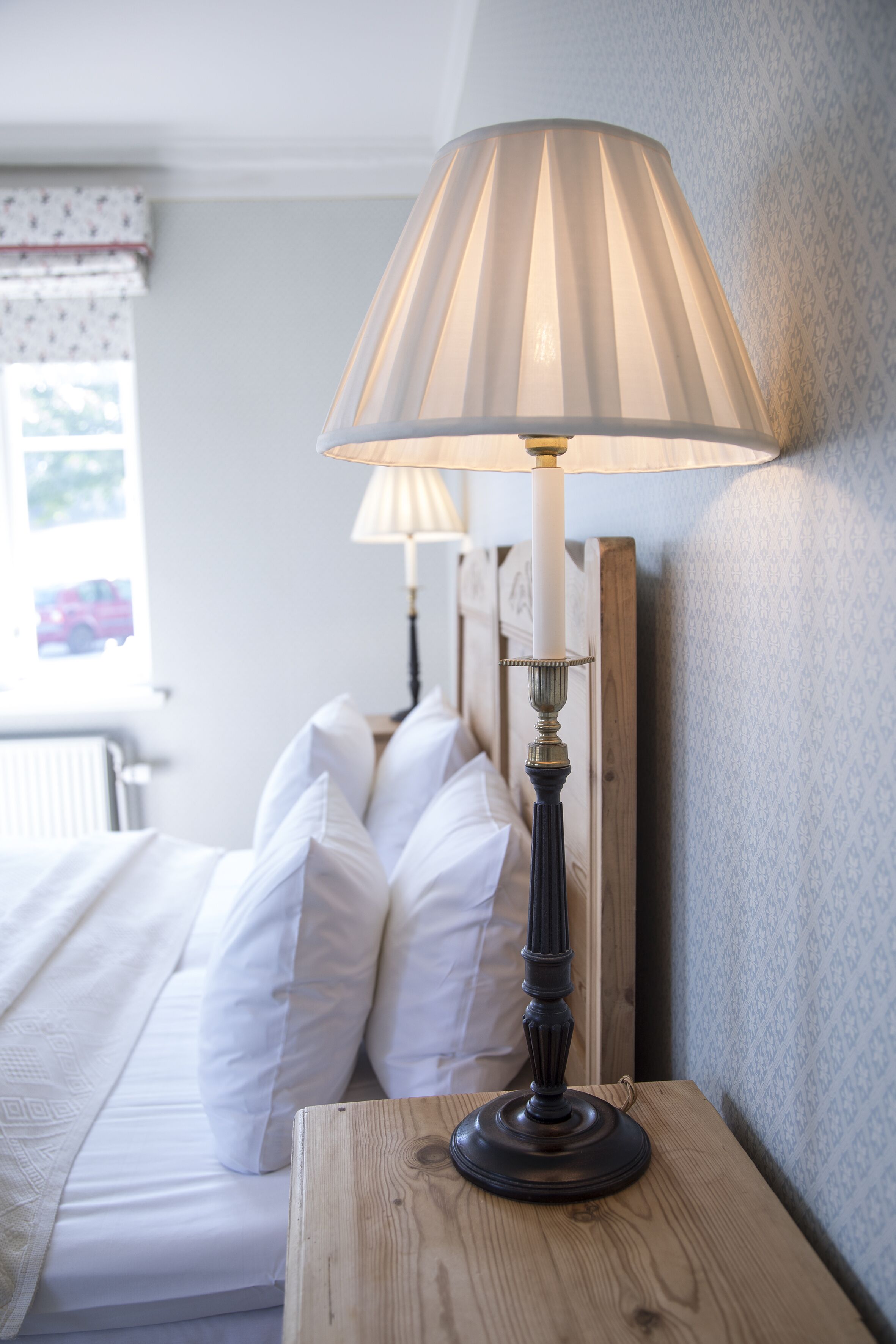 Uma cama com lençóis e almofadas brancos, uma mesa de cabeceira de madeira com abajur, e parede com papel de parede estampado.