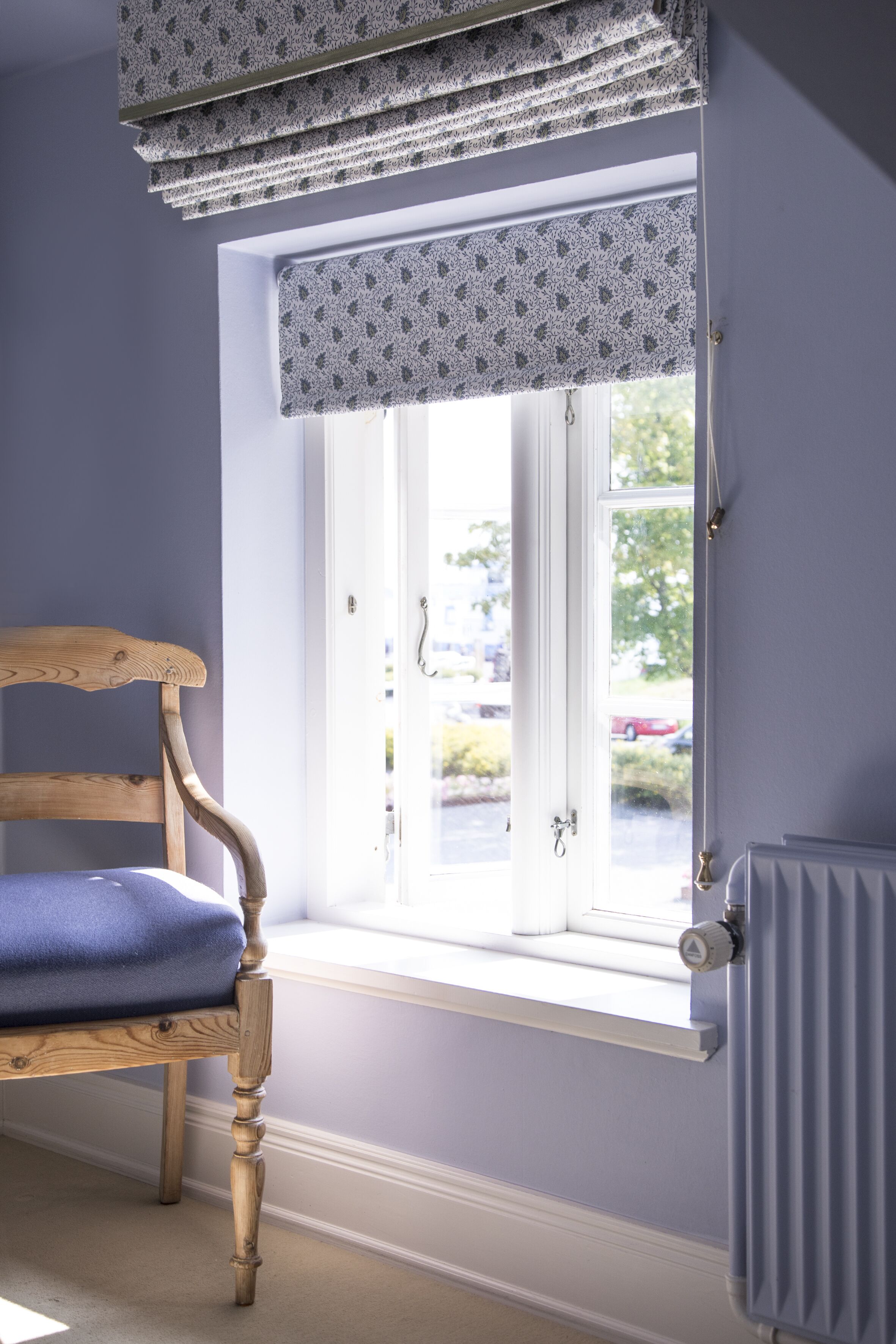 A room with a window covered by patterned valance, a wooden chair with a cushion, and a radiator on the wall.