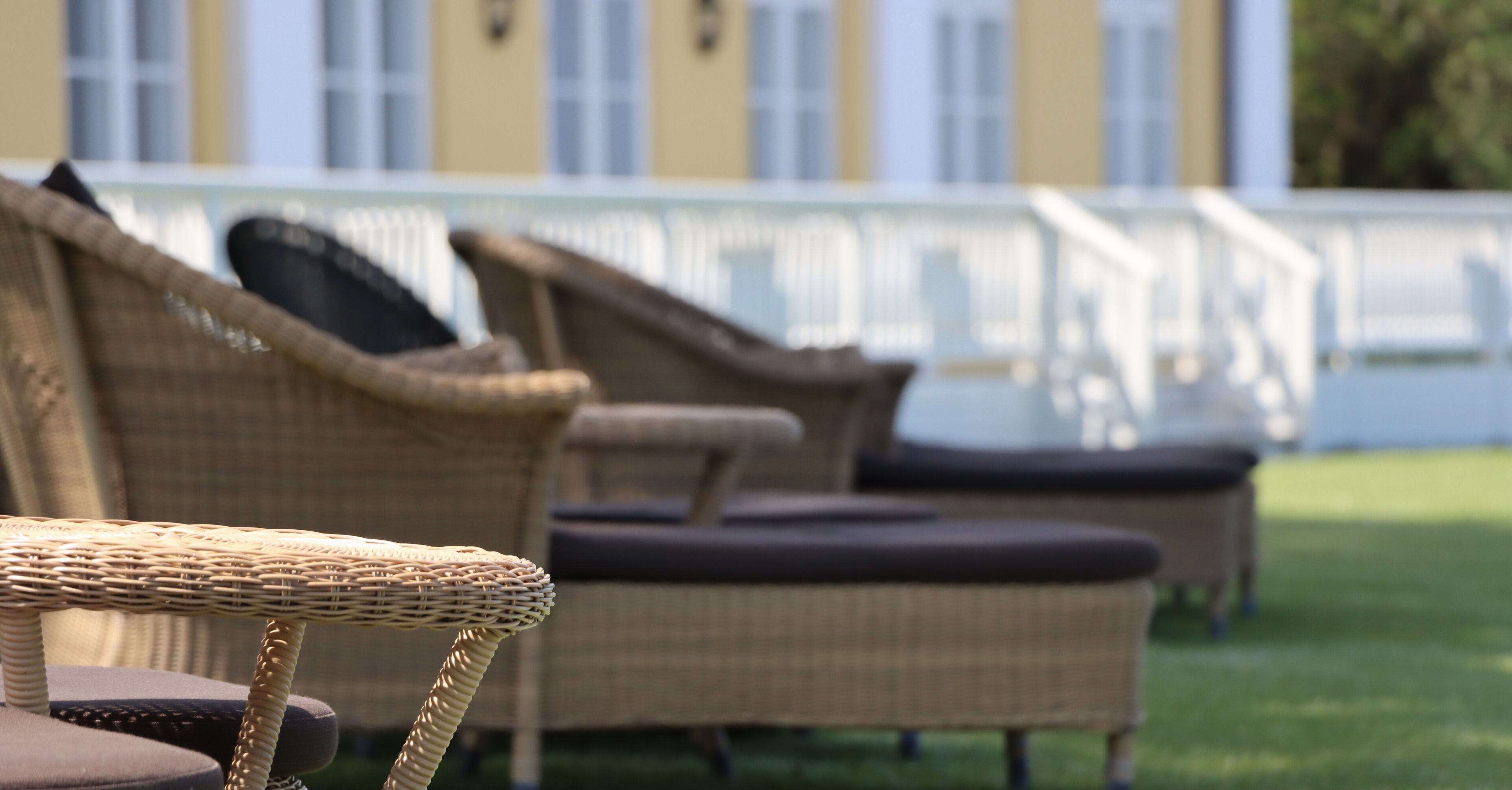 Wicker lounge chairs with cushions, arranged outdoors with a white fence and a building in the background.