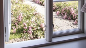 A view through a window with a white frame, leading to a garden with pink flowers and a large tree. - Ballebro Færgekro (Sønderborg)