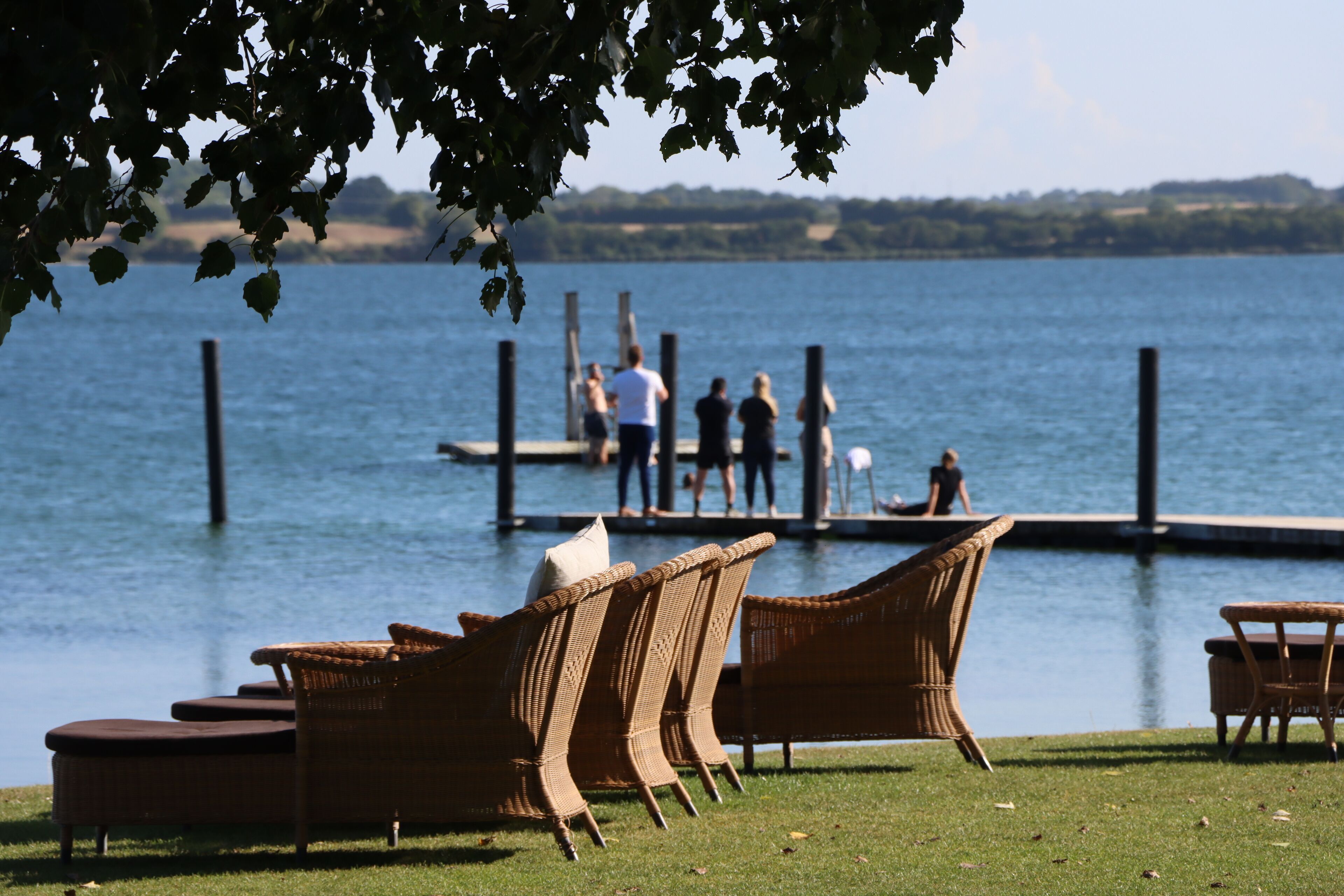 Wicker furniture on a grassy area by a lake, with a dock in the background where people are present.