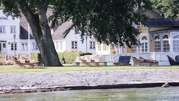 Un hotel frente al agua con terraza al aire libre, un árbol grande y un edificio con techo de paja.