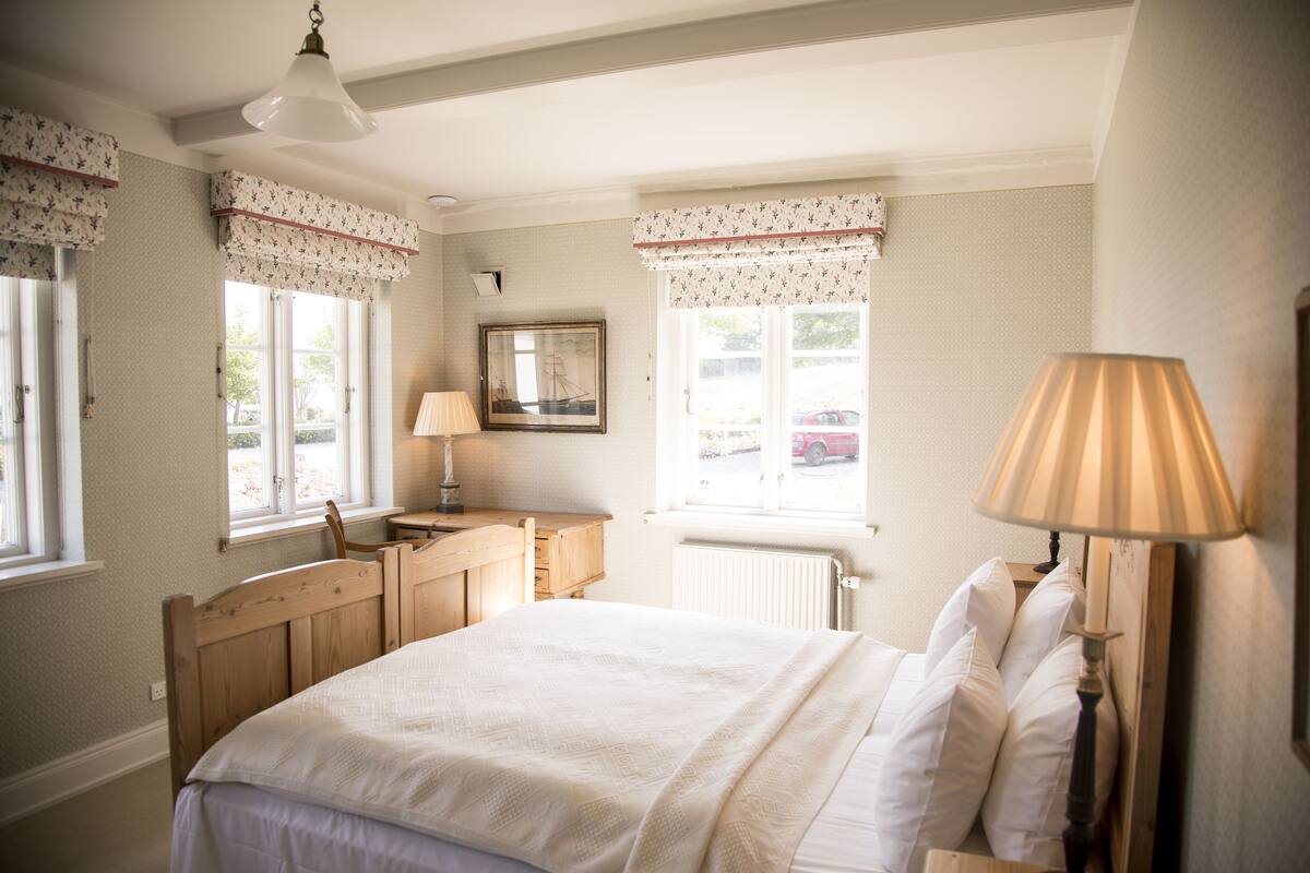 Ein Schlafzimmer mit Bett, Schreibtisch, Stuhl, Lampe und Fenster mit Blick nach draußen.