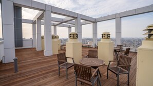 Una terraza en la azotea con suelo de madera, mesas y sillas redondas, y vistas a la ciudad.