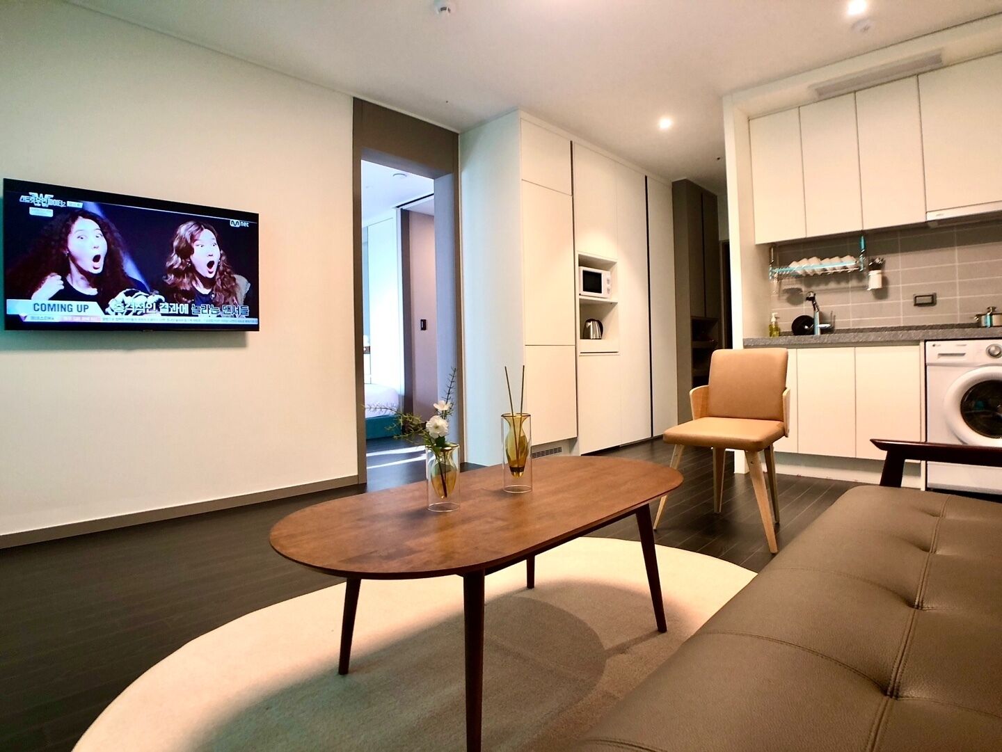 A modern living room with a TV mounted on the wall, a round wooden coffee table, a beige sofa, and a kitchen area with white cabinets and a washing machine.