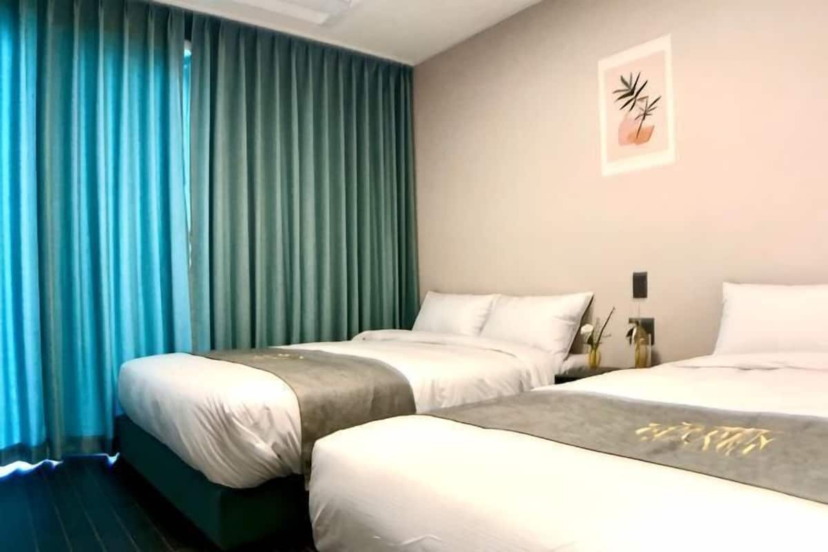 Ett hotellrum med tvÄ sÀngar, ett stort fönster med gardiner, ett konstverk som hÀnger pÄ vÀggen och ett nattduksbord med en vas.